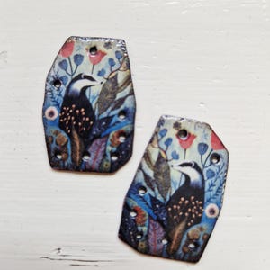 Puede incluir: Dos colgantes decorativos de forma irregular con un diseño temático de la naturaleza. Cada colgante presenta una ilustración de un pájaro con la cabeza en blanco y negro, rodeado de flores y follaje coloridos en tonos azules, rojos y verdes. Los colgantes tienen pequeños agujeros para colgar.