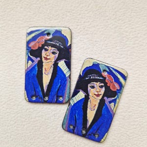 May include: Two rectangular pendants with a vintage-style portrait of a woman in a hat and blue coat. The artwork uses blue, black, red, and yellow. Each pendant has three small holes for attachment, and the overall style is reminiscent of early 20th-century art.