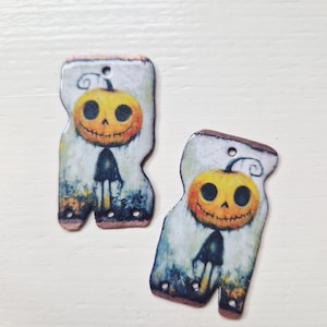 Könnte beinhalten: Zwei Halloween-Holzanhänger. Jeder Anhänger zeigt einen lächelnden Kürbiskopf mit schwarzem Körper. Der Hintergrund hat einen Aquarelleffekt mit gedämpften Farben. Die Anhänger haben oben ein Loch zum Aufhängen.