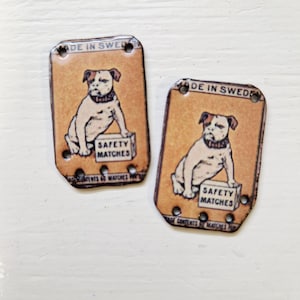Könnte beinhalten: Zwei rechteckige, metallene Streichholzschachtel-Cover im Vintage-Stil. Jedes zeigt eine sitzende Bulldoggen-Illustration über einem Schild mit der Aufschrift "SAFETY MATCHES". Der Text "MADE IN SWEDEN" steht oben.