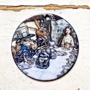May include: A round ceramic ornament featuring a whimsical illustration of the Mad Hatter, the March Hare, and Alice from Alice in Wonderland. The scene depicts a tea party with a teapot, cups, and saucers.