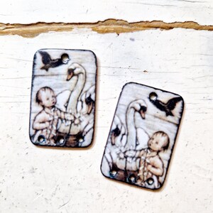 May include: Two rectangular wooden tags with a white background and a printed image of a baby holding onto two swans with a black bird flying overhead. The tags have three holes each.