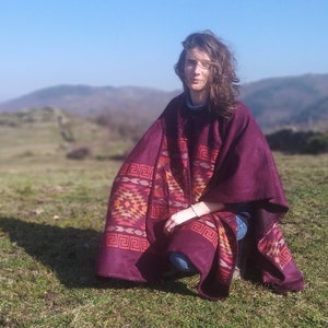Puede incluir: Una mujer con un poncho granate con un patrón geométrico se sienta en una colina cubierta de hierba. El poncho tiene un patrón rojo, naranja y amarillo. El fondo es una vista borrosa de una colina cubierta de hierba con árboles en la distancia.