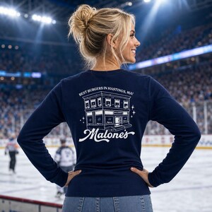 May include: A navy blue long-sleeve shirt featuring a white graphic on the back. The graphic depicts a building with the text "BEST BURGERS IN HASTINGS, MA!" above it and the word "Malone's" in a large script font underneath the building.