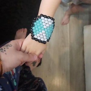 May include: A wide, beaded cuff bracelet in shades of blue, white, and black. The bracelet is worn on a wrist and held by another person's hand. The bracelet is made of small, square beads.