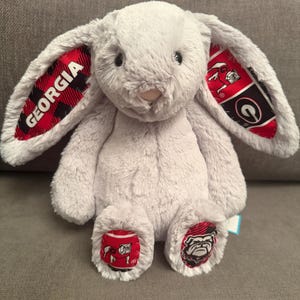 May include: A light gray plush rabbit with long, floppy ears. The inner ears feature a red and black plaid pattern with the word GEORGIA printed on one side and a bulldog logo on the other. The feet have red and black patterned fabric patches.