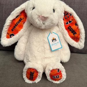 May include: A cream-colored plush rabbit with long ears featuring an orange Syracuse University pattern on the inner lining. The rabbit has a pink nose and a Jellycat London tag attached to its neck. The feet also feature the orange Syracuse pattern.