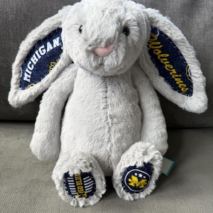 May include: A light gray plush rabbit with long ears featuring dark blue fabric linings printed with the text MICHIGAN and Wolverines. The rabbit has a pink nose and dark eyes. The soles of the feet are dark blue with the text GO BLUE and a Michigan logo.