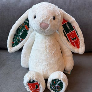 May include: A cream-colored plush rabbit with long, floppy ears. The inner lining of the ears and the soles of the feet feature a green, orange, and white University of Miami logo pattern. The rabbit has a soft, textured fur finish and a pink nose.