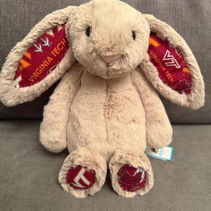 May include: A beige plush rabbit toy with long ears featuring a maroon and orange pattern with the text Virginia Tech and the VT logo. The rabbit has matching maroon and orange patterned fabric on the soles of its feet and a small tag on its side.