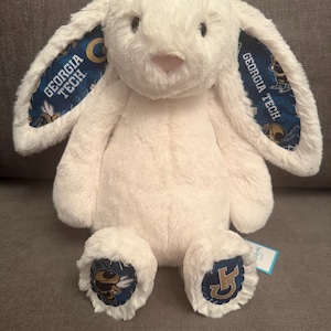 May include: A cream-colored plush rabbit with long, floppy ears. The inner lining of the ears features a blue pattern with the text Georgia Tech and a mascot graphic. The rabbit's feet also display blue fabric patches with the Georgia Tech logo.