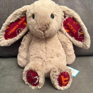 May include: A plush bunny rabbit in a light brown color. The inside of the ears and the soles of the feet feature a maroon fabric with the words "VIRGINIA TECH" and "HOKIES" in gold lettering. The bunny has black eyes and a pink nose.
