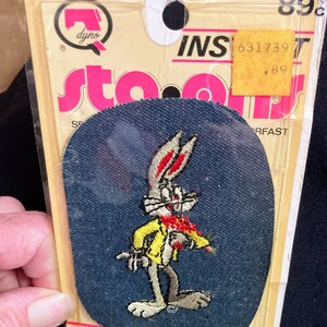 May include: A blue denim iron-on patch featuring Bugs Bunny in a yellow jacket holding a carrot. The patch is packaged on a card with the text "DYNO" and "INS 631739 T .89" printed on it.