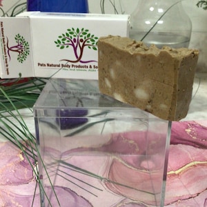 Op de afbeelding: Een rechthoekige zeepblok met een lichtbruine kleur en witte vlekken, rustend op een doorzichtige acrylbox. Een witte doos met de tekst "Pats Natural Body Products & Soap" en een boomlogo is zichtbaar. Een blauwe vaas en een glazen vaas staan op de achtergrond.