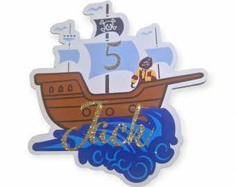 May include: A blue and brown pirate ship cake topper with a pirate figure on the deck, a skull and crossbones flag, and the number 5. The name Jack is written in gold glitter on the waves below the ship.