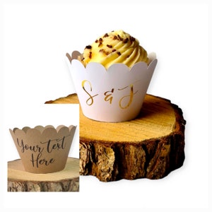 May include: White cupcake liner with scalloped edges and gold foil lettering that says "S & J". The liner is on a wooden slice. A second cupcake liner is shown in the image, this one is brown with the text "Your Text Here" in black.