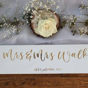 May include: White wooden sign with gold lettering that reads "Mrs & Mrs Walker" and the date "19th September 2022".