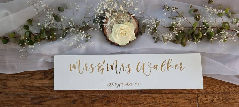 May include: White wooden sign with gold lettering that reads "Mrs & Mrs Walker" and the date "19th September 2022".