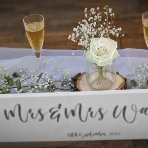 May include: White wooden sign with the text "Mrs & Mrs Walker" and the date "28th September 2022" in black calligraphy. Two champagne flutes with champagne are on a white tablecloth with baby's breath flowers.