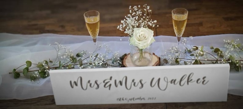 May include: White wooden sign with the text "Mrs & Mrs Walker" and the date "28th September 2022" in black calligraphy. Two champagne flutes with champagne are on a white tablecloth with baby's breath flowers.