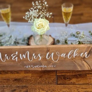 May include: A rustic wooden sign with the text "Mr & Mrs Walker 19th September 2022" in white lettering. The sign is on a white tablecloth with greenery and flowers.