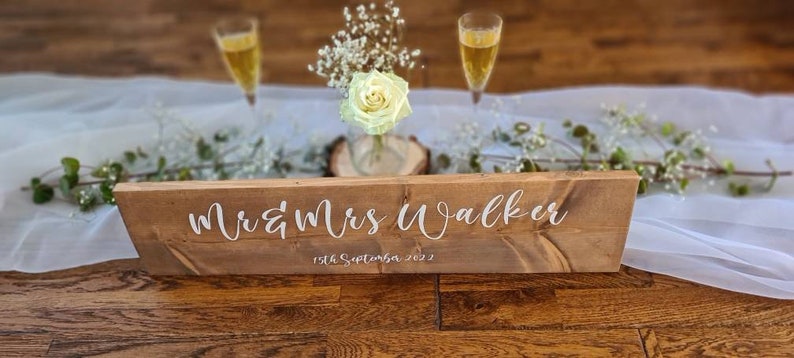 May include: A rustic wooden sign with the text "Mr & Mrs Walker 19th September 2022" in white lettering. The sign is on a white tablecloth with greenery and flowers.
