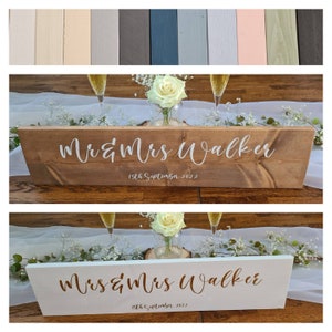 May include: Two wooden wedding signs with the text "Mr & Mrs Walker" and "15th September 2022" in white and gold lettering. The signs are on a white table runner with white flowers and champagne glasses.