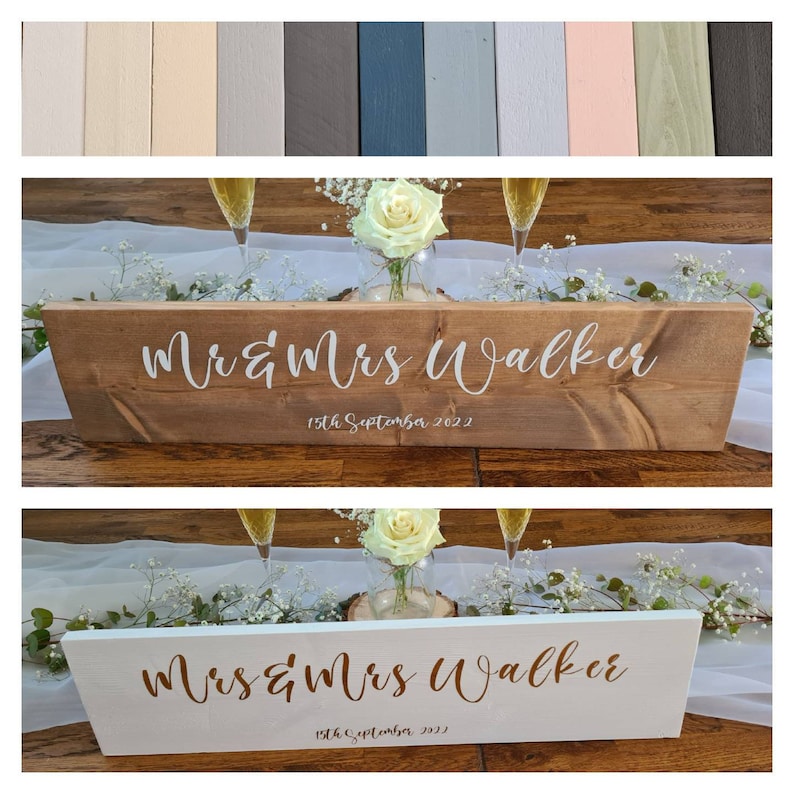 May include: Two wooden wedding signs with the text "Mr & Mrs Walker" and "15th September 2022" in white and gold lettering. The signs are on a white table runner with white flowers and champagne glasses.