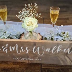 May include: A rustic wooden sign with the text "Mr & Mrs Walker" and the date "13th September 2023" in white lettering. The sign is decorated with white flowers and two champagne flutes.