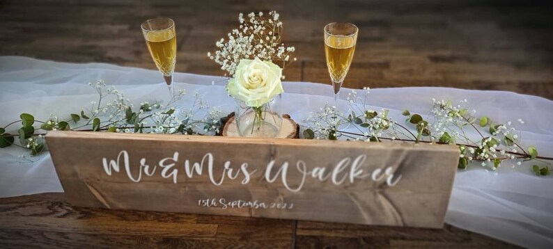 May include: A rustic wooden sign with the text "Mr & Mrs Walker" and the date "13th September 2023" in white lettering. The sign is decorated with white flowers and two champagne flutes.
