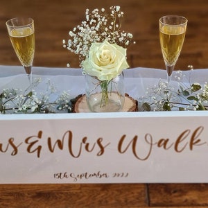 May include: White wooden sign with the text "Mrs & Mrs Walker" and the date "15th September 2023" in gold lettering. The sign is decorated with white flowers and two champagne flutes.