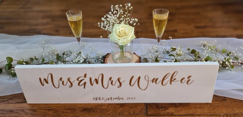 May include: White wooden sign with the text "Mrs & Mrs Walker" and the date "15th September 2023" in gold lettering. The sign is decorated with white flowers and two champagne flutes.