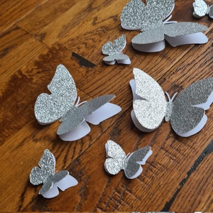 May include: A set of eight silver glitter butterflies of varying sizes. The butterflies are made of paper and have a 3D effect.