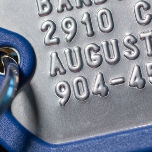 K&ouml;nnte beinhalten: Nahaufnahme eines silbernen Metallschildes mit dem gepr&auml;gten Text "2910 AUGUSTA 904-455". Das Schild ist an einem blauen Kunststoffring befestigt.