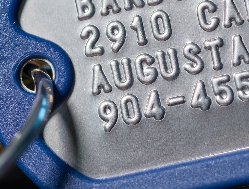 K&ouml;nnte beinhalten: Nahaufnahme eines silbernen Metallschildes mit dem gepr&auml;gten Text "2910 AUGUSTA 904-455". Das Schild ist an einem blauen Kunststoffring befestigt.