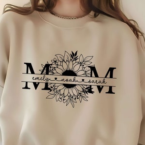 May include: A beige sweatshirt with a black graphic design of the word "MOM" with a sunflower and floral design. The names "emily", "noah", and "sarah" are written below the sunflower.
