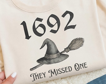May include: Cream-colored sweatshirt with a black graphic featuring the number "1692", a witch's hat, and a broom. The text "THEY MISSED ONE" is printed below the graphic. The sweatshirt is made of a soft material.