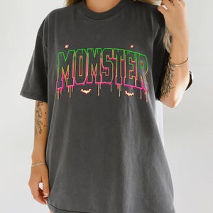 May include: Dark gray t-shirt with the word "MONSTER" in a neon green and pink gradient font. The word has dripping accents and small bat silhouettes. The shirt is a casual, oversized fit.
