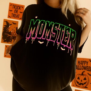 May include: Black t-shirt with the word "MOMSTER" in a neon green and pink gradient font. The design includes dripping lines and small bat silhouettes. Halloween-themed decorations are visible in the background.