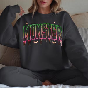 May include: A dark gray sweatshirt with the word "MOMSTER" in a neon green and pink gradient font. The design includes dripping lines and small bat silhouettes, creating a Halloween theme. The sweatshirt is part of a loungewear set.