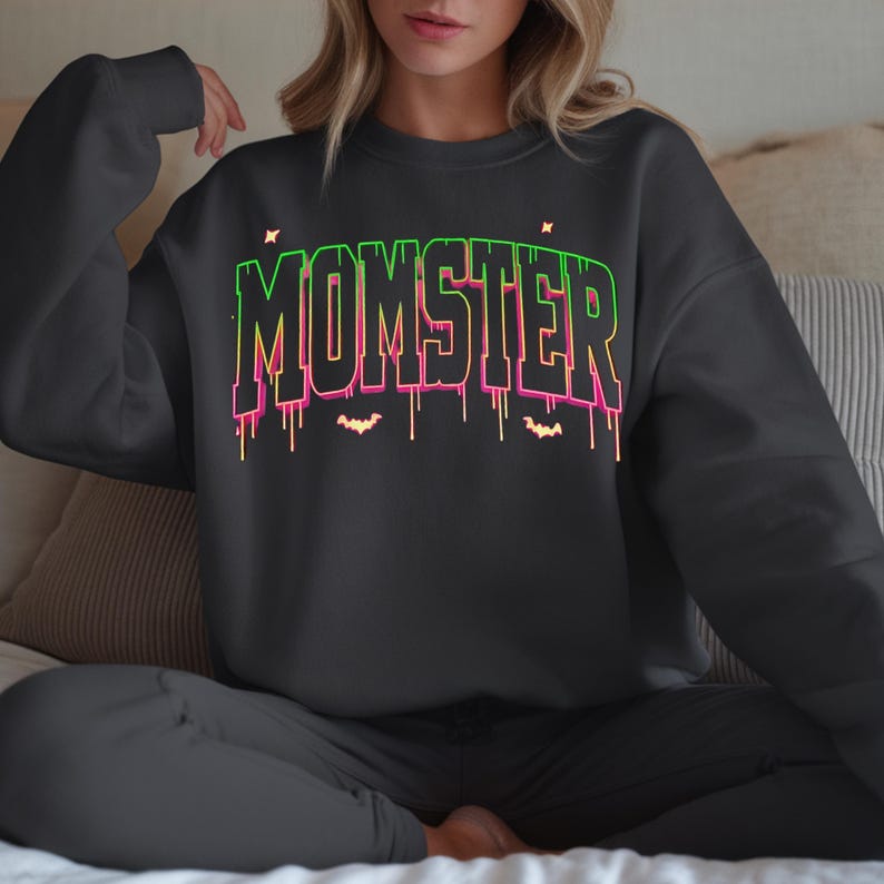 May include: A dark gray sweatshirt with the word "MOMSTER" in a neon green and pink gradient font. The design includes dripping lines and small bat silhouettes, creating a Halloween theme. The sweatshirt is part of a loungewear set.