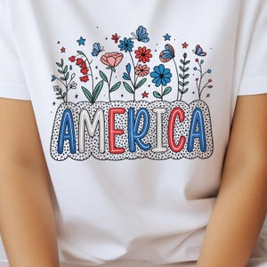 Peut inclure: T-shirt blanc avec un motif de fleurs et de papillons rouges, blancs et bleus avec le mot "AMERICA" dans une police de caract&egrave;res stylis&eacute;e.