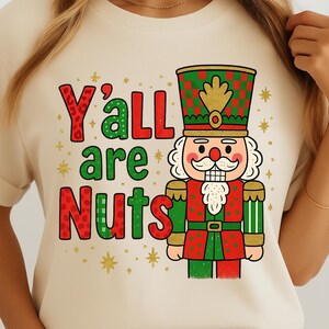 May include: A cream-coloured t-shirt featuring the phrase "Y'all are Nuts" in red and green, with a cartoon nutcracker soldier in red, green, and gold. Gold stars are scattered around the text and image, creating a festive design.