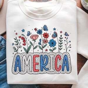 Peut inclure: Sweat-shirt blanc avec un motif graphique de fleurs et d'&eacute;toiles rouges, blanches et bleues avec le mot "AMERICA" dans une police color&eacute;e.