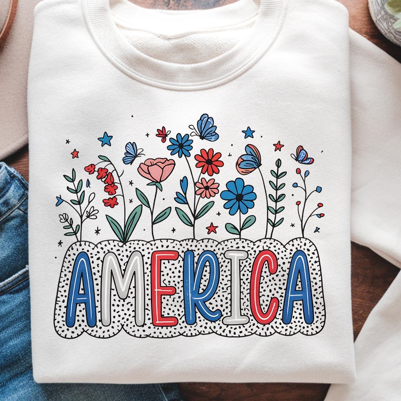 Peut inclure: Sweat-shirt blanc avec un motif graphique de fleurs et d'&eacute;toiles rouges, blanches et bleues avec le mot "AMERICA" dans une police color&eacute;e.