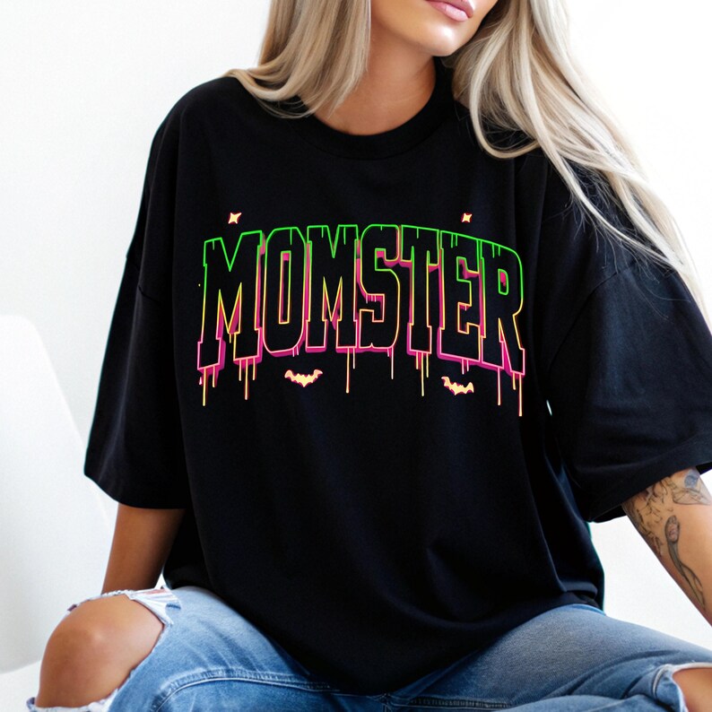 May include: Black t-shirt with the word "MOMSTER" in neon green and pink, with dripping accents and bat silhouettes. The shirt is a relaxed fit, perfect for Halloween or a spooky theme.