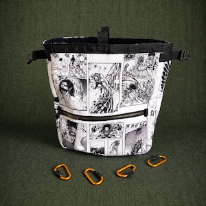 May include: A white and black comic-style chalk bag with a black drawstring closure and a zippered pocket. The bag features a black strap and buckle closure. Four orange carabiners are at the base of the bag. The bag is on a green surface.