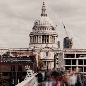 Puede incluir: Una imagen borrosa de personas caminando por un puente frente a la Catedral de San Pablo en Londres, Inglaterra. La catedral es un edificio grande y blanco con una cúpula y una alta aguja. El cielo está nublado y gris.