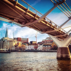 May include: A view of the Millennium Bridge in London, England, with the Shard skyscraper in the background. The bridge is made of steel and glass and has a modern design. The sky is blue and there are clouds in the sky.