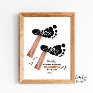 May include: Framed print with a light wood frame. The artwork features two black footprints forming hammers, with nails below. Text reads "Daddy, we love building memories with you! Love,"